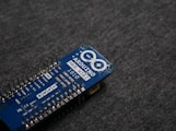 A blue Arduino MKR WiFi 1010 microcontroller board placed on a gray fabric surface. The board features numerous metal pins along one side and is marked with white text identifying its model and specifications.