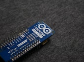 A blue Arduino MKR WiFi 1010 microcontroller board placed on a gray fabric surface. The board features numerous metal pins along one side and is marked with white text identifying its model and specifications.