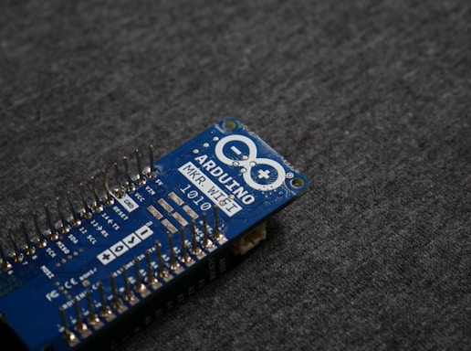 A blue Arduino MKR WiFi 1010 microcontroller board placed on a gray fabric surface. The board features numerous metal pins along one side and is marked with white text identifying its model and specifications.
