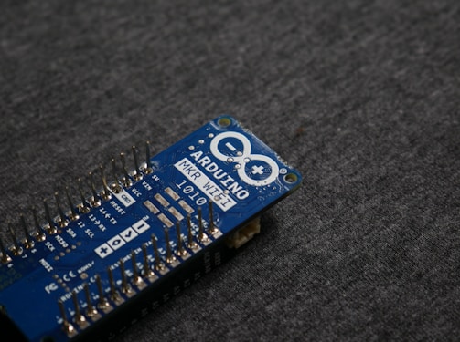 A blue Arduino MKR WiFi 1010 microcontroller board placed on a gray fabric surface. The board features numerous metal pins along one side and is marked with white text identifying its model and specifications.