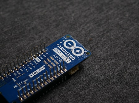 A blue Arduino MKR WiFi 1010 microcontroller board placed on a gray fabric surface. The board features numerous metal pins along one side and is marked with white text identifying its model and specifications.