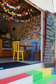 A small café with a colorful and inviting atmosphere. There are multicolored chairs at a wooden table, vibrant decorations hanging from the ceiling, and numerous handwritten notes covering the walls. A chalkboard menu is displayed outside, listing items such as pizzas, pastas, and beverages. The steps leading to the entrance are painted in bright rainbow colors.