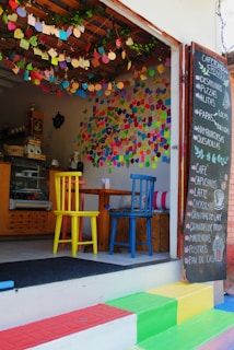 A small café with a colorful and inviting atmosphere. There are multicolored chairs at a wooden table, vibrant decorations hanging from the ceiling, and numerous handwritten notes covering the walls. A chalkboard menu is displayed outside, listing items such as pizzas, pastas, and beverages. The steps leading to the entrance are painted in bright rainbow colors.