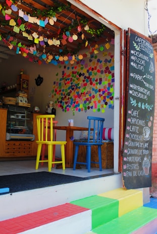 A small café with a colorful and inviting atmosphere. There are multicolored chairs at a wooden table, vibrant decorations hanging from the ceiling, and numerous handwritten notes covering the walls. A chalkboard menu is displayed outside, listing items such as pizzas, pastas, and beverages. The steps leading to the entrance are painted in bright rainbow colors.