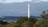 A tall smokestack rises above a dense forested area with a backdrop of cloud-covered skies. In the distance, a small city with numerous buildings is visible. Lush green foliage surrounds the base of the smokestack, contrasting with the urban landscape further away.