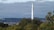 A tall smokestack rises above a dense forested area with a backdrop of cloud-covered skies. In the distance, a small city with numerous buildings is visible. Lush green foliage surrounds the base of the smokestack, contrasting with the urban landscape further away.