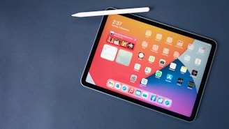 A tablet with a stylus resting on its surface displays a colorful home screen filled with various app icons. The screen shows widgets for time, music, and weather updates, featuring a modern and sleek design. The device is placed on a dark, flat surface.