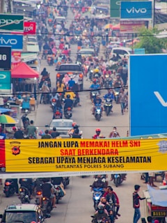 A busy street filled with motorcycles and cars, with a prominent yellow sign in the foreground urging people to wear helmets for safety. Advertisements and shop signs line the sides of the street, and a dense crowd of riders can be seen in the background.