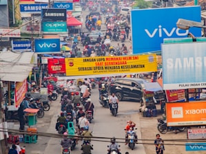 The image captures a busy street scene bustling with motorcycles and people. Numerous billboards and signs featuring technology brands like Samsung, Vivo, Oppo, and Xiaomi are prominently displayed. There is a large yellow banner across the street with text reminding riders to wear helmets for safety. The street is crowded, with people walking and riding motorcycles, indicating a lively and chaotic urban environment.