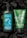 Two Bath & Body Works aromatherapy products featuring stress relief properties are placed against a dark background. The left product is a green bottle of moisturizing body lotion infused with eucalyptus and tea, adorned with water droplets. On the right is a green tube of invigorating body scrub with eucalyptus and spearmint. Both products are surrounded by pieces of black charcoal.