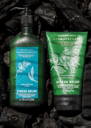 Two Bath & Body Works aromatherapy products featuring stress relief properties are placed against a dark background. The left product is a green bottle of moisturizing body lotion infused with eucalyptus and tea, adorned with water droplets. On the right is a green tube of invigorating body scrub with eucalyptus and spearmint. Both products are surrounded by pieces of black charcoal.