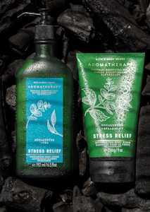 Two Bath & Body Works aromatherapy products featuring stress relief properties are placed against a dark background. The left product is a green bottle of moisturizing body lotion infused with eucalyptus and tea, adorned with water droplets. On the right is a green tube of invigorating body scrub with eucalyptus and spearmint. Both products are surrounded by pieces of black charcoal.