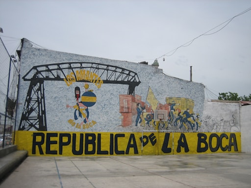 A large mural painted on a wall, featuring colorful images and text. It includes a stylized figure holding a globe with a painterly, abstract background. Several smaller figures appear to be engaged in lively activities, adjacent to a depiction of an industrial structure. Bold text in yellow and black spells out 'Los Amantes de La Boca Republica de La Boca' across the mural.