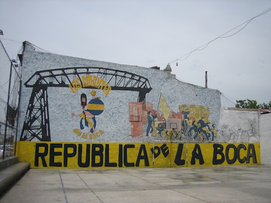 A large mural painted on a wall, featuring colorful images and text. It includes a stylized figure holding a globe with a painterly, abstract background. Several smaller figures appear to be engaged in lively activities, adjacent to a depiction of an industrial structure. Bold text in yellow and black spells out 'Los Amantes de La Boca Republica de La Boca' across the mural.