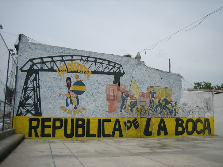 A large mural painted on a wall, featuring colorful images and text. It includes a stylized figure holding a globe with a painterly, abstract background. Several smaller figures appear to be engaged in lively activities, adjacent to a depiction of an industrial structure. Bold text in yellow and black spells out 'Los Amantes de La Boca Republica de La Boca' across the mural.