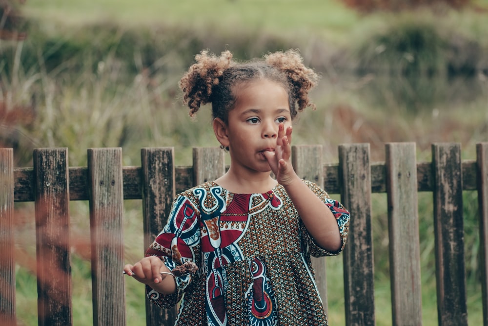 girl in blue and white floral dress standing near brown wooden fence during daytime