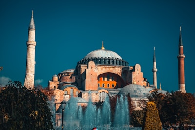 A grand architectural structure with a large central dome flanked by several smaller domes and four minarets. The building is surrounded by lush greenery and a fountain in the foreground.