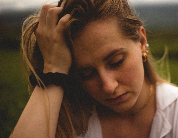 A person with light brown hair appears distressed or deep in thought, with their hand resting on their head. They are wearing a white shirt and gold earrings, with a black hairband around their wrist. The background is blurred out, suggesting an outdoor setting.