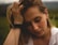 A person with light brown hair appears distressed or deep in thought, with their hand resting on their head. They are wearing a white shirt and gold earrings, with a black hairband around their wrist. The background is blurred out, suggesting an outdoor setting.