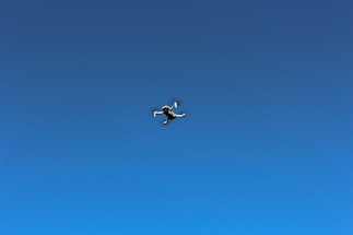 A smiling child wearing pilot goggles, looking up at a small drone flying overhead against a clear blue sky.