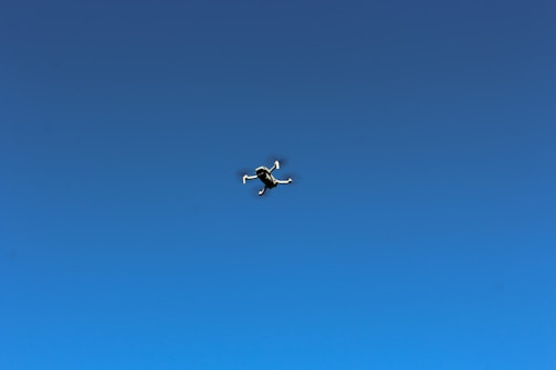 A smiling child wearing pilot goggles, looking up at a small drone flying overhead against a clear blue sky.