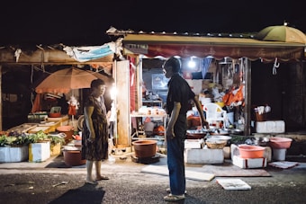 A night market scene shows two people standing in front of a small shop filled with various goods. The surrounding area is cluttered with baskets, boxes, and fresh produce, illuminated by bright lights under an awning. The shop appears weathered and busy, with an umbrella and a plastic chair enhancing the lively market atmosphere.