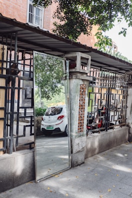 A mirror is placed in an outdoor setting, reflecting a white car and surrounding greenery. The structure around the mirror is composed of a gray frame with black bars and red brick accents. A neighboring building's brick wall with windows and leafy tree branches add to the scene.