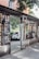 A mirror is placed in an outdoor setting, reflecting a white car and surrounding greenery. The structure around the mirror is composed of a gray frame with black bars and red brick accents. A neighboring building's brick wall with windows and leafy tree branches add to the scene.