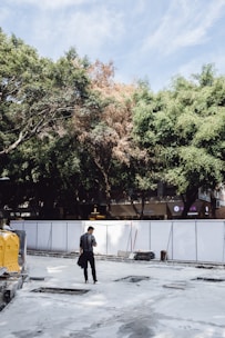 A solitary person stands on a construction site surrounded by white barriers. The ground appears unfinished, with some exposed sections. In the background, lush trees with varying shades of green and some brown cover a portion of the sky. Shops or buildings are partially visible behind the trees.