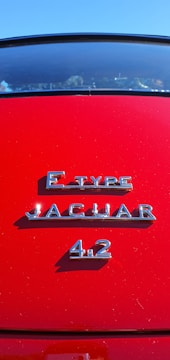 A close-up of a classic car's rear, showcasing polished chrome lettering indicating it's a Jaguar E-Type 4.2 against a vibrant red background.