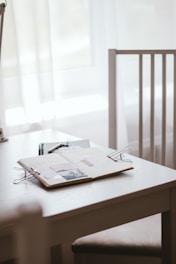A workspace setting is depicted with a white table and a wooden chair. On the table lies an open notebook with photos and a paper clip, alongside a closed black notebook and a pen. Soft natural light filters through white curtains, creating a serene atmosphere.