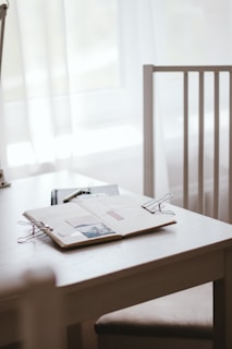 A workspace setting is depicted with a white table and a wooden chair. On the table lies an open notebook with photos and a paper clip, alongside a closed black notebook and a pen. Soft natural light filters through white curtains, creating a serene atmosphere.