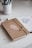 A brown notebook with a white botanical design on its cover lies closed on a white desk. The notebook is secured with a piece of twine. Next to it, there is an open sketchbook with an illustration of a teapot, and a white cup containing various pens and pencils.