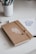 A brown notebook with a white botanical design on its cover lies closed on a white desk. The notebook is secured with a piece of twine. Next to it, there is an open sketchbook with an illustration of a teapot, and a white cup containing various pens and pencils.