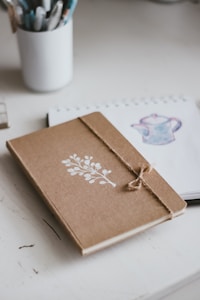 A brown notebook with a white botanical design on its cover lies closed on a white desk. The notebook is secured with a piece of twine. Next to it, there is an open sketchbook with an illustration of a teapot, and a white cup containing various pens and pencils.