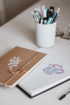 A neatly arranged desk includes a brown notebook with a simple white floral design and a spiral-bound sketchbook open to a page featuring a teapot sketch. Several pens and pencils are organized in a white cup, adding to the creative atmosphere.