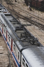 Aerial view of a train on railway tracks, showcasing the overhead electric lines above the train. The train has a metallic exterior with red, blue, and white stripes. The tracks diverge into multiple directions, and a rail yard with other rail cars is visible in the background.