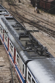 Aerial view of a train on railway tracks, showcasing the overhead electric lines above the train. The train has a metallic exterior with red, blue, and white stripes. The tracks diverge into multiple directions, and a rail yard with other rail cars is visible in the background.