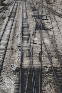 A complex network of railway tracks interwoven with multiple switches and crossings, showing an intricate pattern of parallel steel rails on wooden ties. The surface is covered with gravel and stones, with mechanical components and signs visible alongside the tracks.