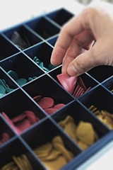 A hand is selecting a pink game piece from a grid-like container filled with various colored game pieces. The container is divided into multiple compartments, each holding pieces of different colors including pink, green, and yellow.