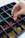 A hand is selecting a pink game piece from a grid-like container filled with various colored game pieces. The container is divided into multiple compartments, each holding pieces of different colors including pink, green, and yellow.
