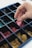 A hand is selecting a pink game piece from a grid-like container filled with various colored game pieces. The container is divided into multiple compartments, each holding pieces of different colors including pink, green, and yellow.