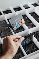 A hand is selecting a card from a grid of small compartments. The card displays three colorful guitar picks in black, blue, pink, and yellow. The background contains several organized compartments holding similar cards. The overall arrangement suggests a meticulous organization, possibly for display or storage.