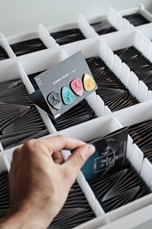 A hand is selecting a card from a grid of small compartments. The card displays three colorful guitar picks in black, blue, pink, and yellow. The background contains several organized compartments holding similar cards. The overall arrangement suggests a meticulous organization, possibly for display or storage.