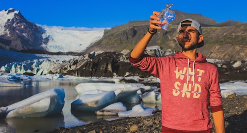 A man is standing in front of a glacier, holding up a piece of ice or crystal towards the sky. He wears a red hoodie and a backward cap. The landscape includes large ice formations and rocky mountains with a clear blue sky above.