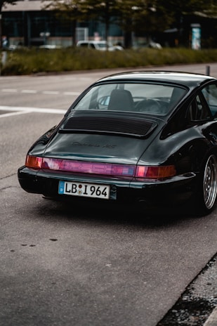 A vintage Porsche 911 Carrera RS parked on a city street, showcasing its sleek rear design with a distinctive license plate. The dark green color of the car contrasts with the urban surroundings, including the pavement and greenery in the background.
