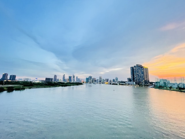 A panoramic view of a city skyline along a river with modern high-rise buildings lining the waterfront. The sky displays a gradient from blue to a warm orange hue as the sun sets, casting reflections on the water.
