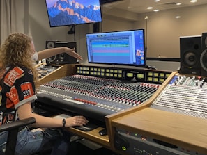 A person with curly hair is seated at an audio mixing console in a recording studio. The mixing board is filled with numerous knobs and sliders. The person is pointing towards a computer screen displaying digital audio tracks. Two large speakers and a smaller control panel are positioned nearby. The room's lighting is subtle, and a curtain is partially visible in the background, along with wall-mounted monitors showing a mountain landscape wallpaper.