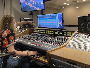 A person with curly hair is seated at an audio mixing console in a recording studio. The mixing board is filled with numerous knobs and sliders. The person is pointing towards a computer screen displaying digital audio tracks. Two large speakers and a smaller control panel are positioned nearby. The room's lighting is subtle, and a curtain is partially visible in the background, along with wall-mounted monitors showing a mountain landscape wallpaper.