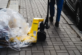 A person pushes a yellow pallet jack along a paved sidewalk. The pallet jack is wrapped in plastic, and the ground is made up of rectangular gray bricks. Part of a parked car is visible on the right side of the image.